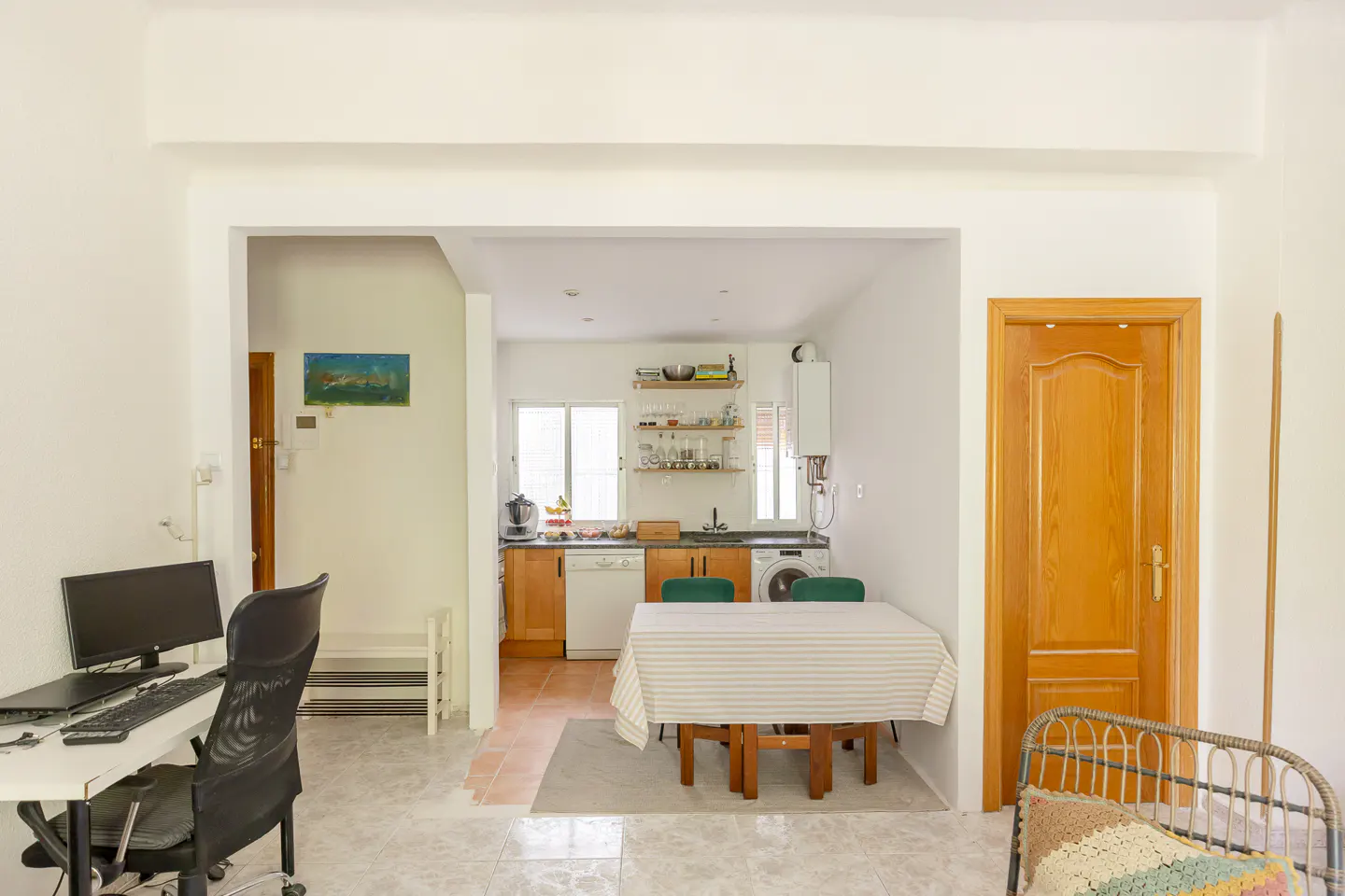 Open-concept home interior with a kitchen, dining table, and office area. The kitchen has wood cabinets and white appliances.