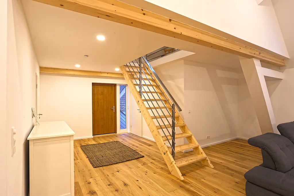 Bright, modern interior with wooden floors, white walls, and exposed beams. A wooden staircase with metal railings leads to an upper level. A brown door and white dresser are visible.