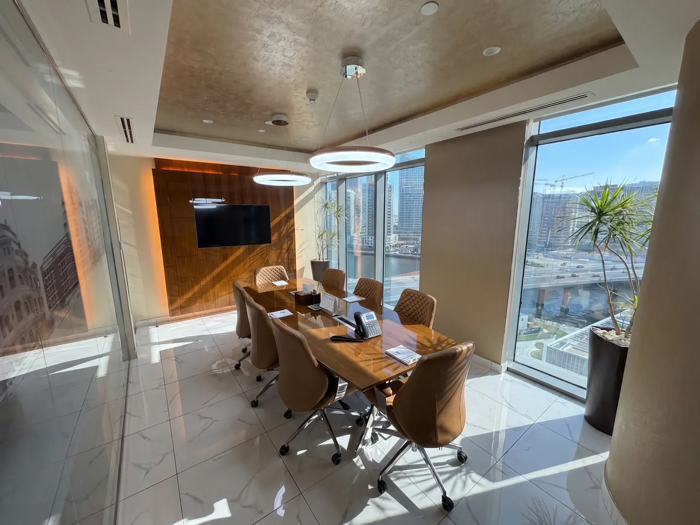 Bright conference room with a long wood table, tan chairs, and city views through large windows. Modern lighting fixtures hang above.
