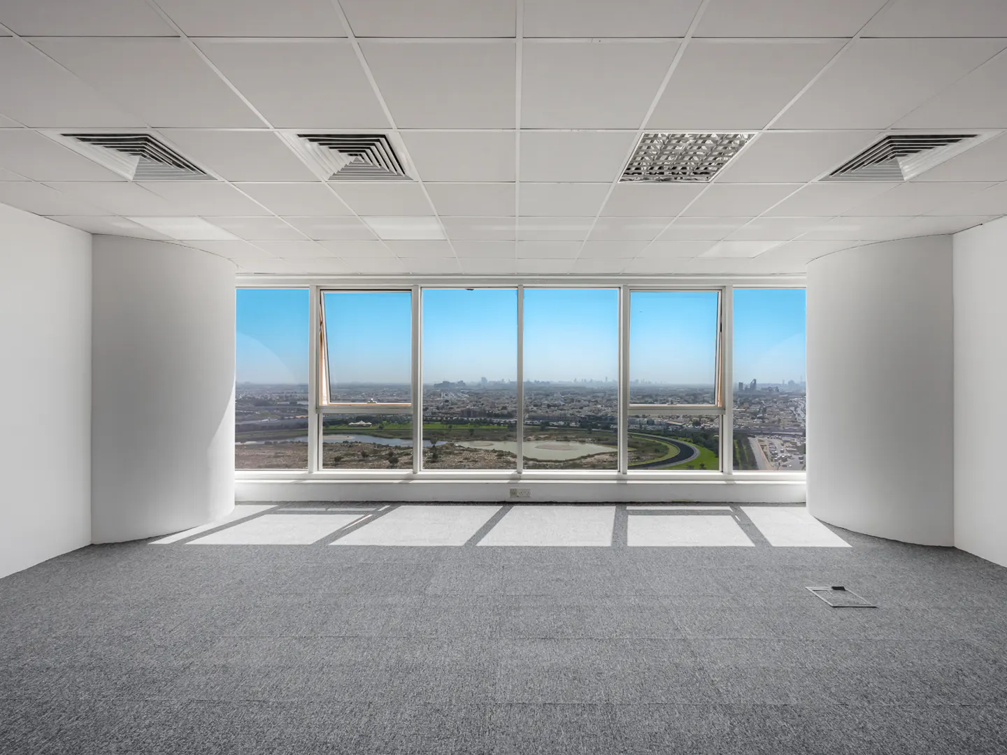 Empty office space with gray carpet, white walls, and large windows showing a city skyline under a blue sky.