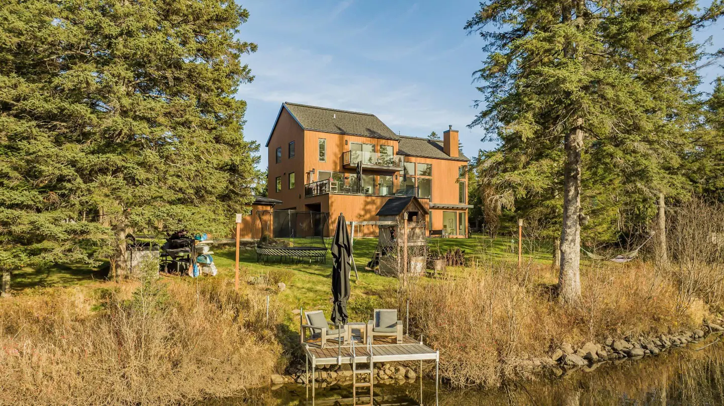 Waterfront property with a dock and chairs in front of a modern, orange house surrounded by tall green trees.