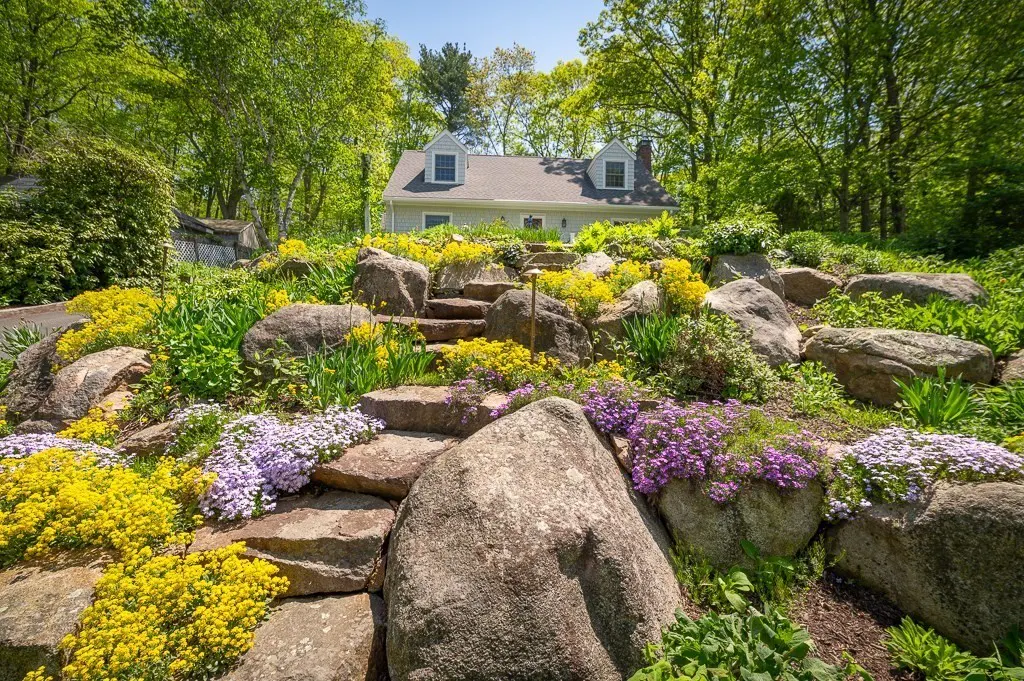 A house sits atop a hill with stone steps and colorful flowers. The house is light green with a gray roof and surrounded by green trees.