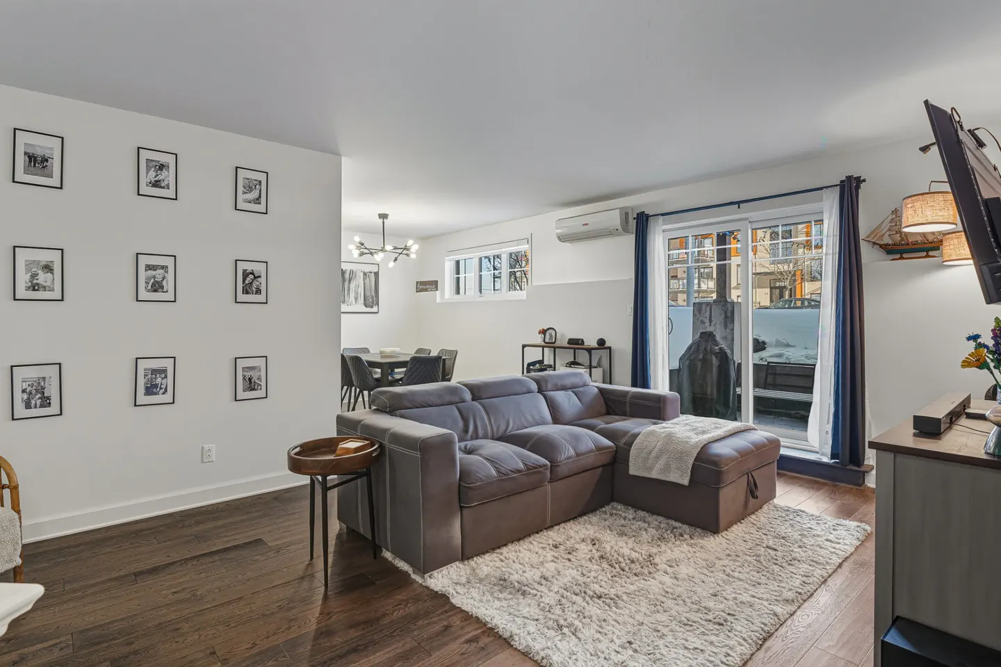 Living room with a gray sofa, white rug, and dark wood floors. A wall of framed photos and a dining area are visible. Sliding glass doors lead to a patio.