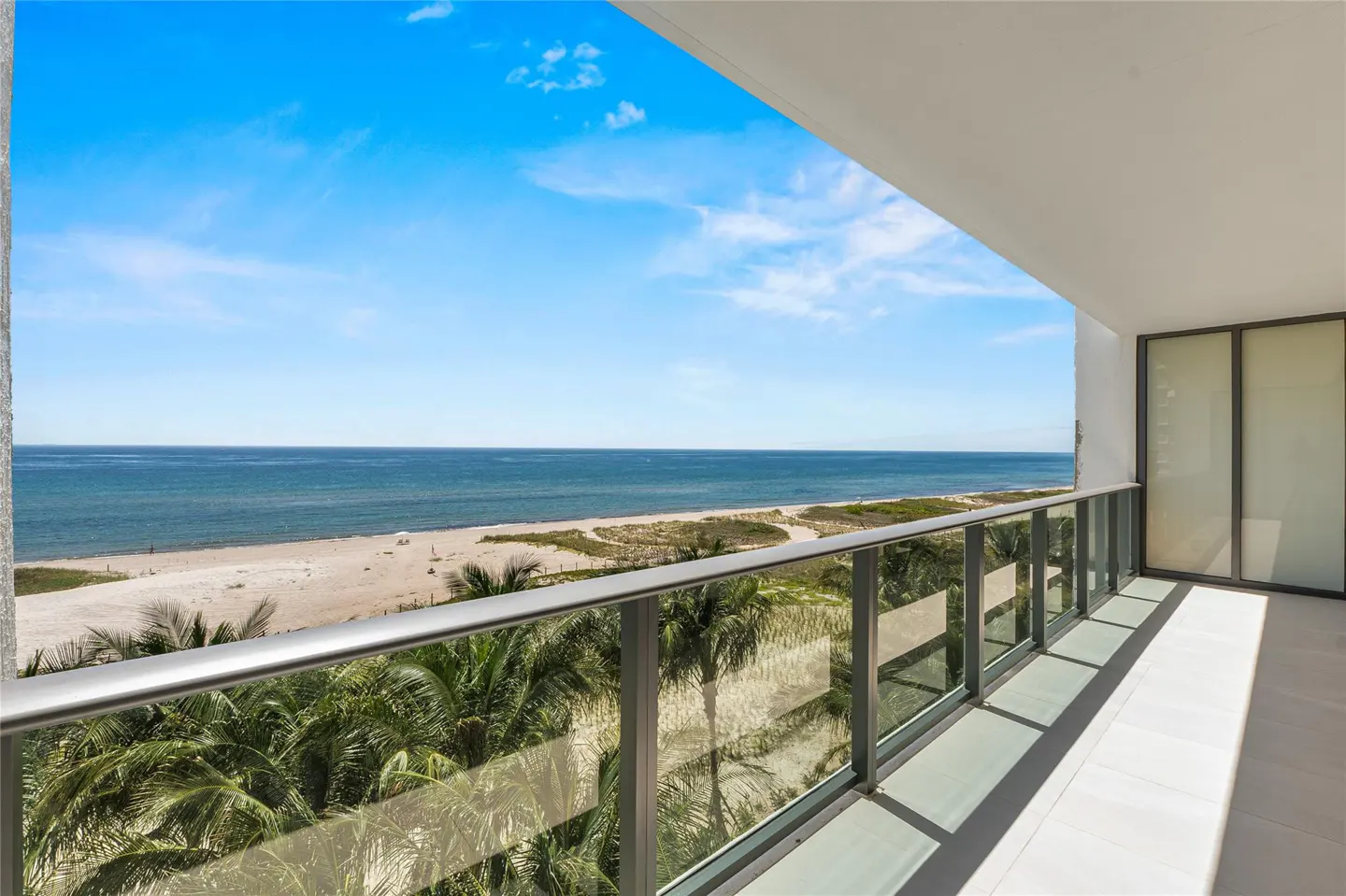 Balcony view of ocean and beach. Glass railing, palm trees, and blue sky.