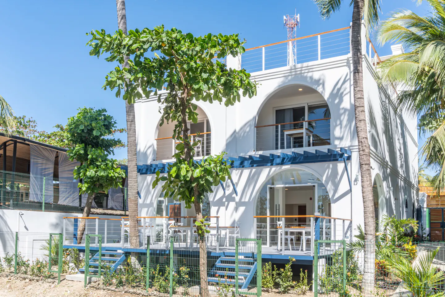 Two-story white building with arched windows and blue trim, surrounded by trees and greenery. Balconies with white furniture.