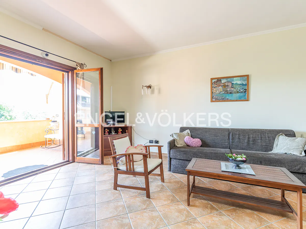 Living room with a gray sofa, wooden chair, and coffee table. Balcony access with open glass doors. Tiled floor. Painting on the wall.