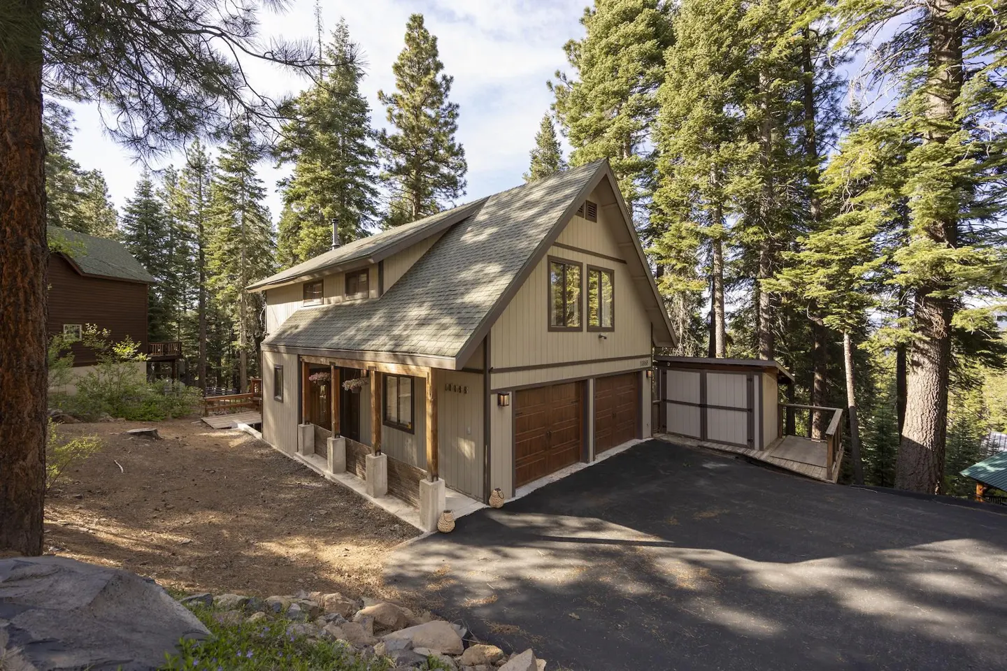 Tan two-story house with a green roof, brown trim, and a two-car garage, surrounded by tall green trees.
