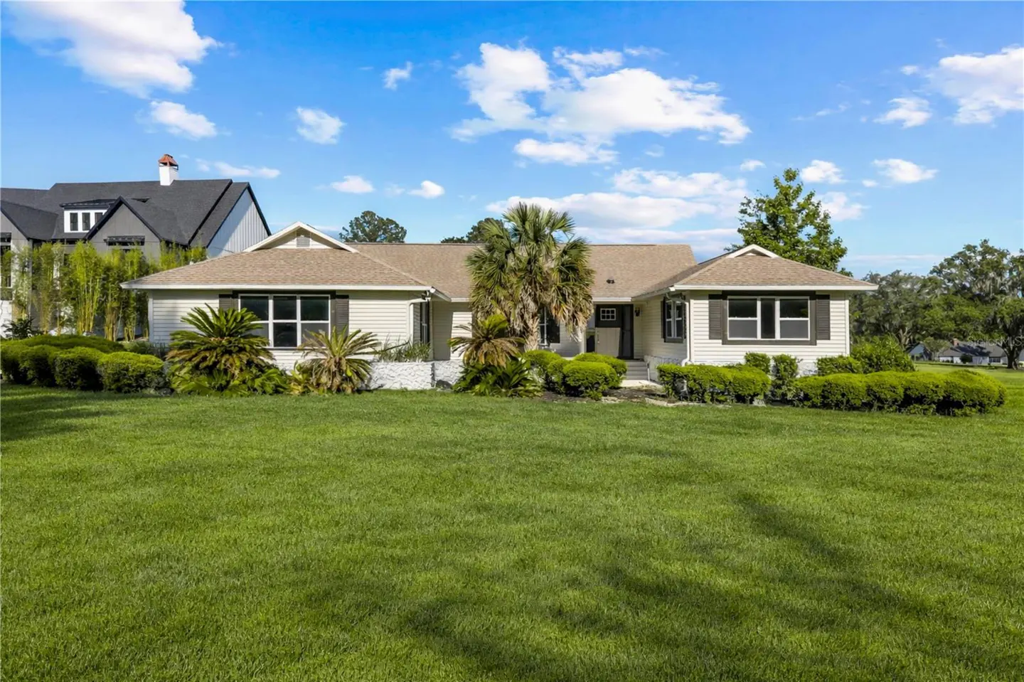 A single-story, light-yellow house with a brown roof, dark shutters, and a large green lawn. Palm trees and bushes surround the house.