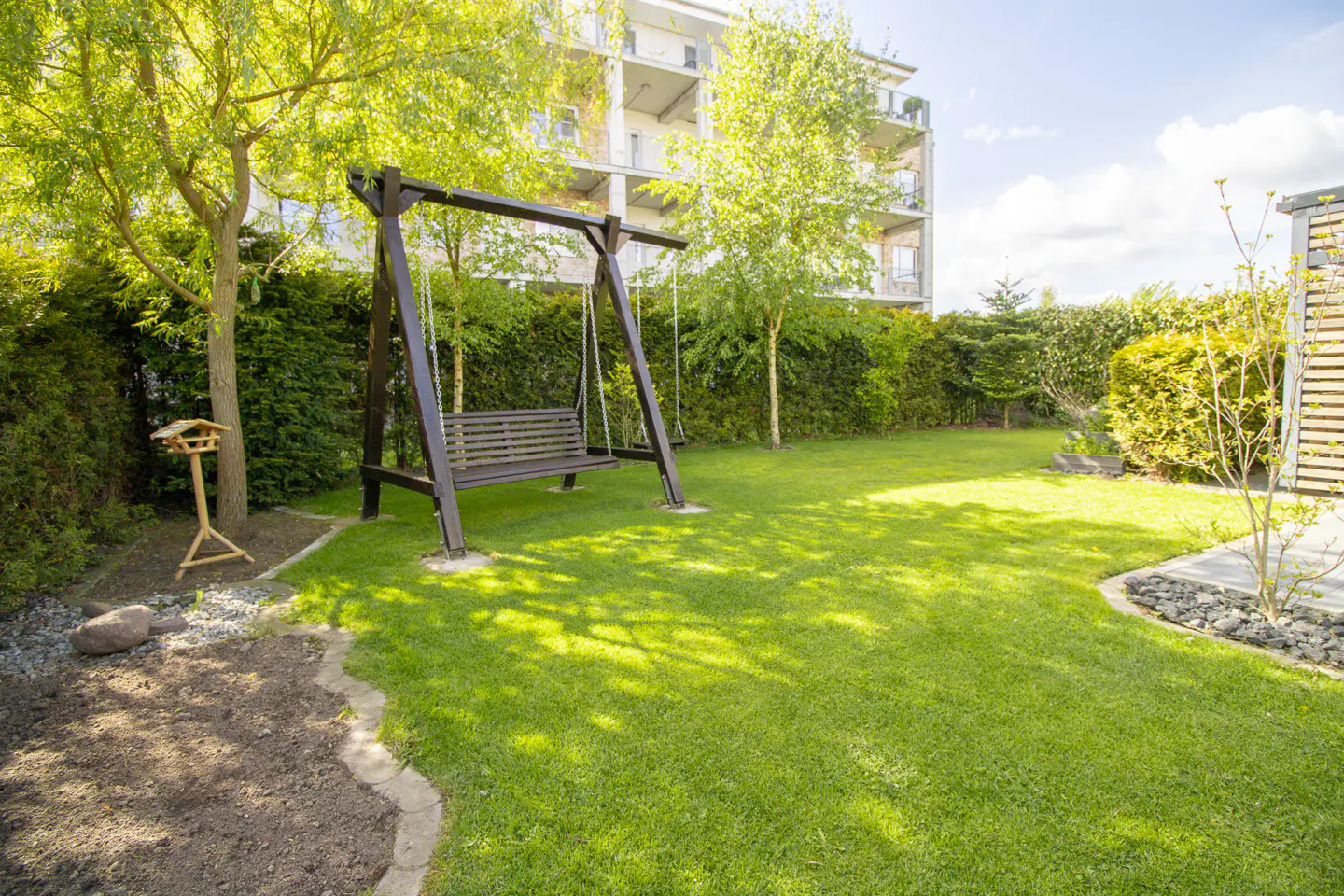 A backyard with a wooden swing set, green grass, trees, and a bird feeder. A building is visible in the background.