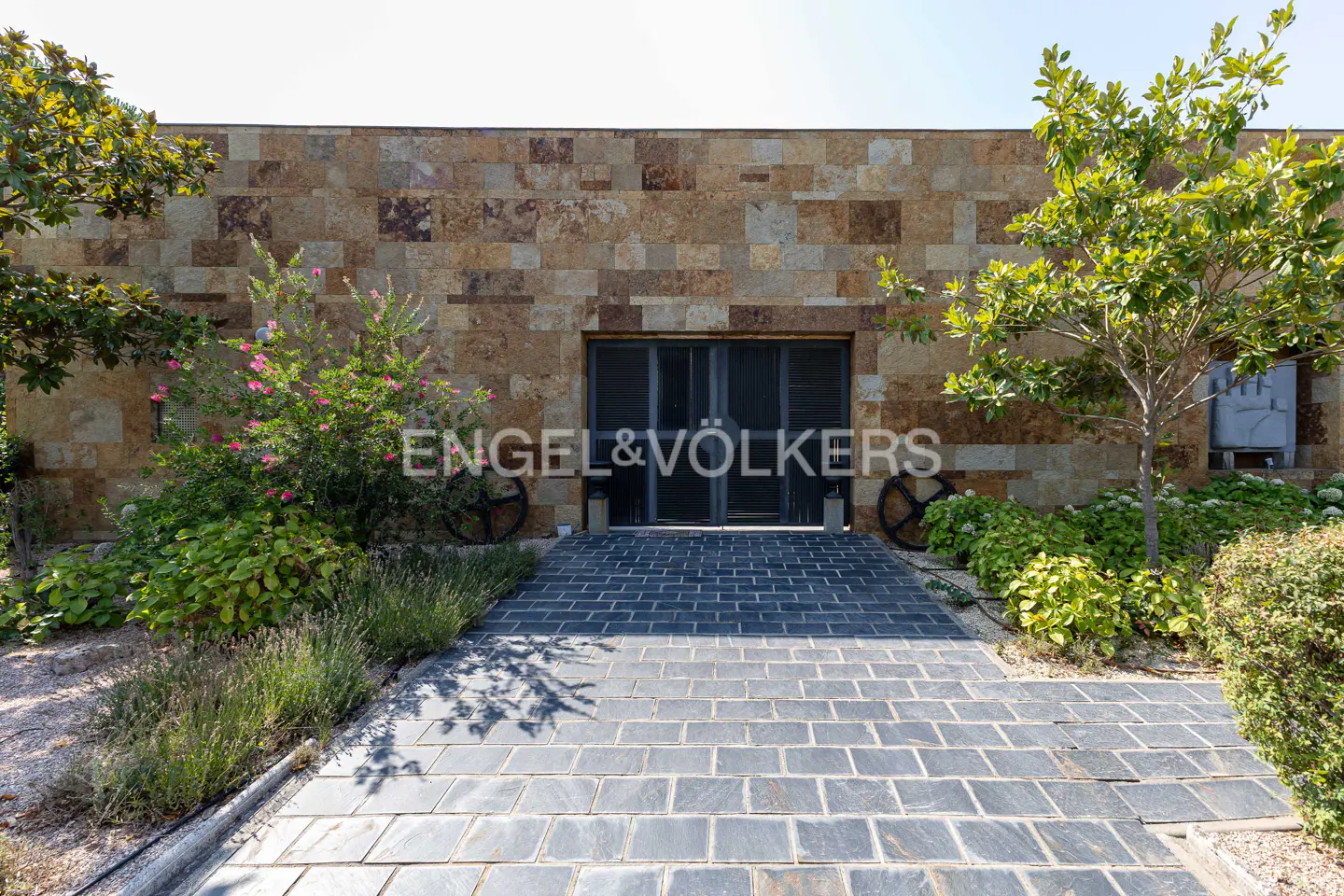 A stone-tiled path leads to a modern entrance with dark glass doors set in a textured, multi-toned stone wall.