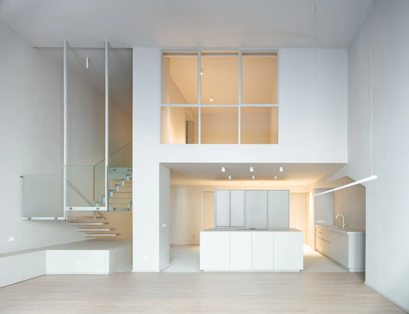 Bright, modern interior with white walls, light wood floors, and a minimalist kitchen. Stairs with glass railings lead to a loft area with a large window.