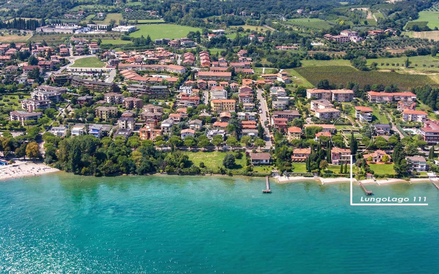 Aerial view of Lungolago 111, a waterfront property with a dock, nestled among houses and green trees near a turquoise lake.