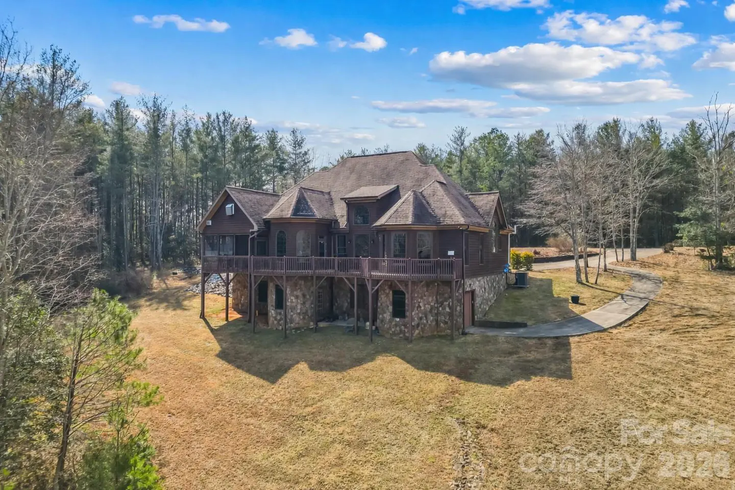 A large, brown, two-story house with a stone foundation and a wooden deck, surrounded by trees and a grassy lawn.