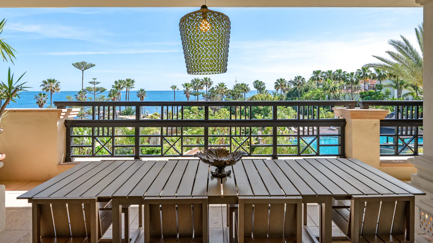 Balcony view with a wooden table and chairs, overlooking palm trees, the ocean, and a blue sky. A decorative light fixture hangs above the table.