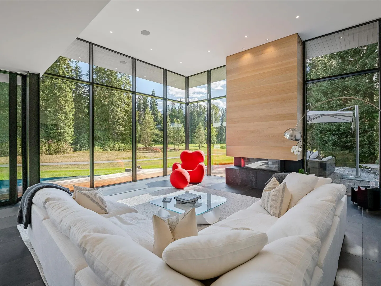 Bright living room with floor-to-ceiling windows, a white sectional sofa, a red chair, and a modern fireplace. Outside is a green lawn and trees.