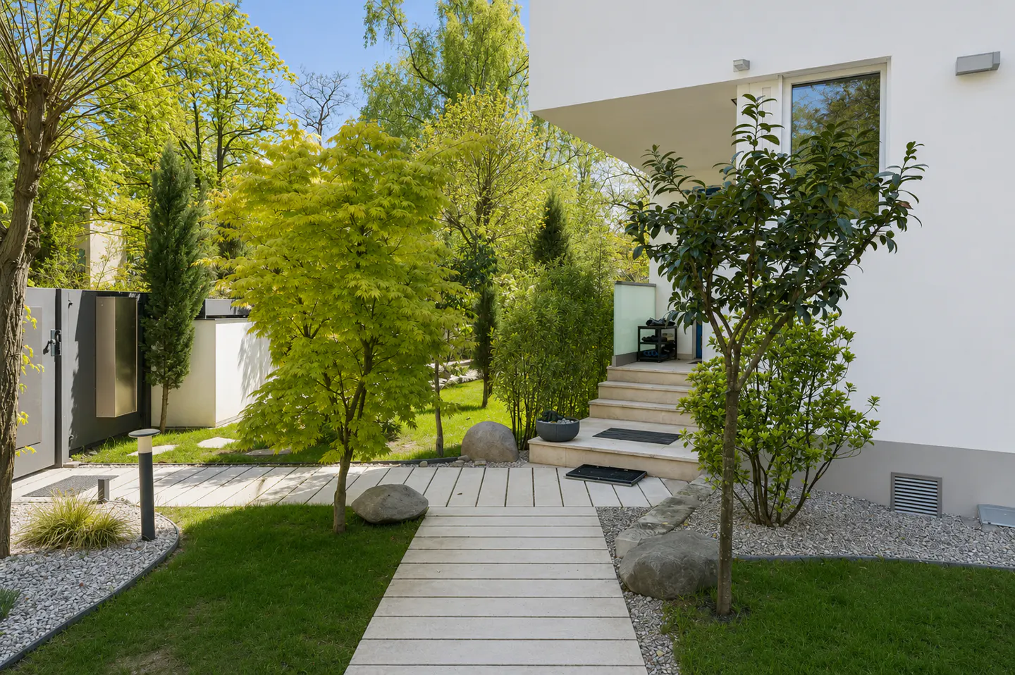 Landscaped front yard with a stone walkway leading to the entrance of a modern white house with green trees and grass.