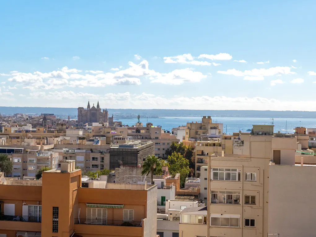 Cityscape view of Palma de Mallorca, Spain, with beige buildings, the sea, and the cathedral under a blue sky.