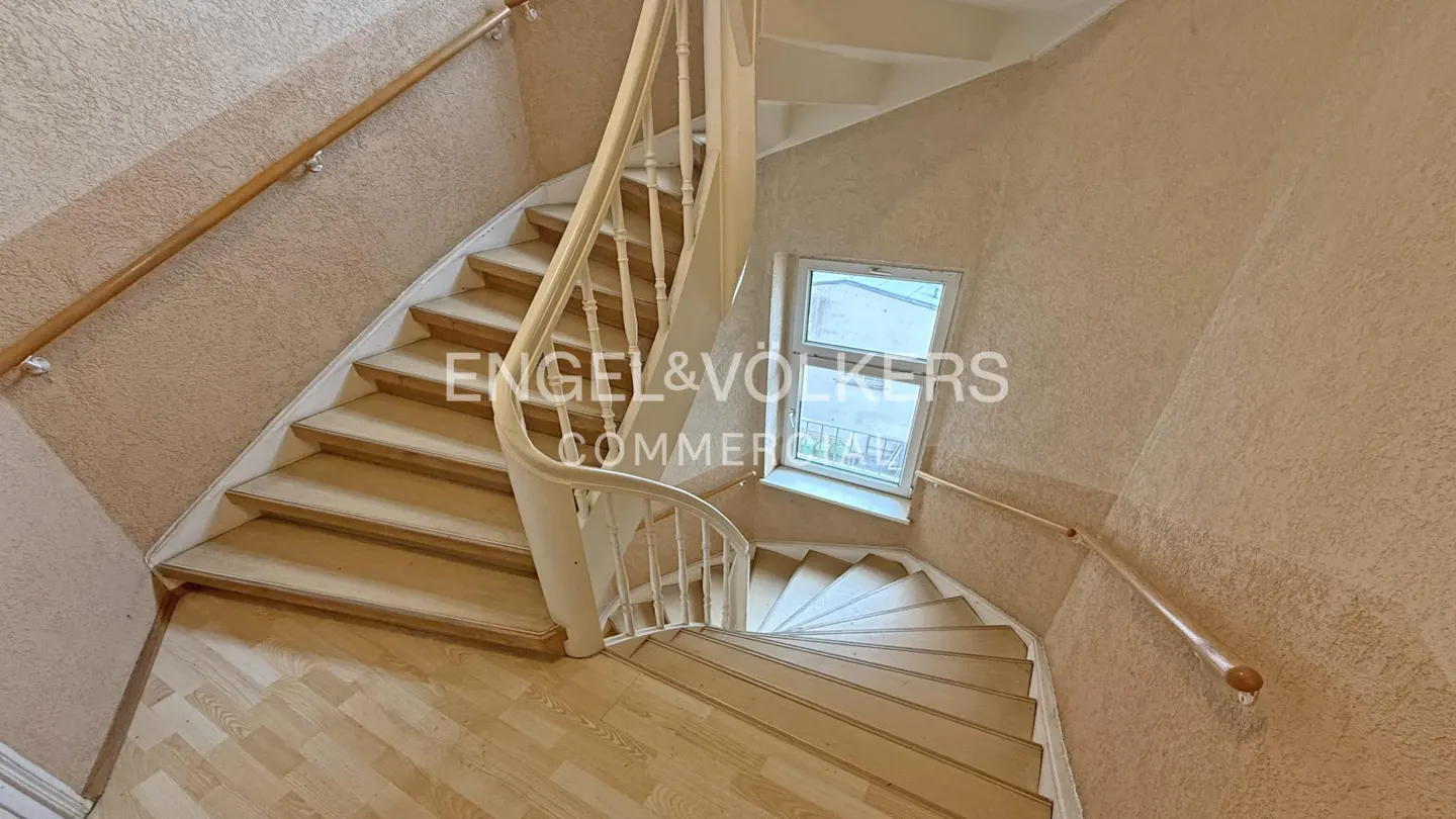 Interior view of a winding staircase with white banister and wooden handrail, light wood floors, and a window.