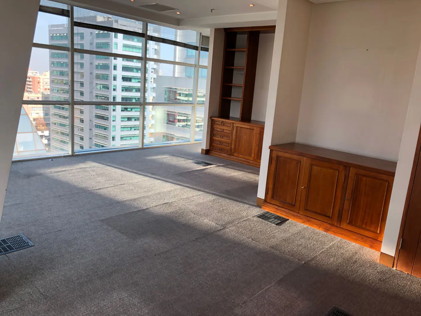 Empty office space with gray carpet, wooden cabinets, and a large window overlooking city buildings.