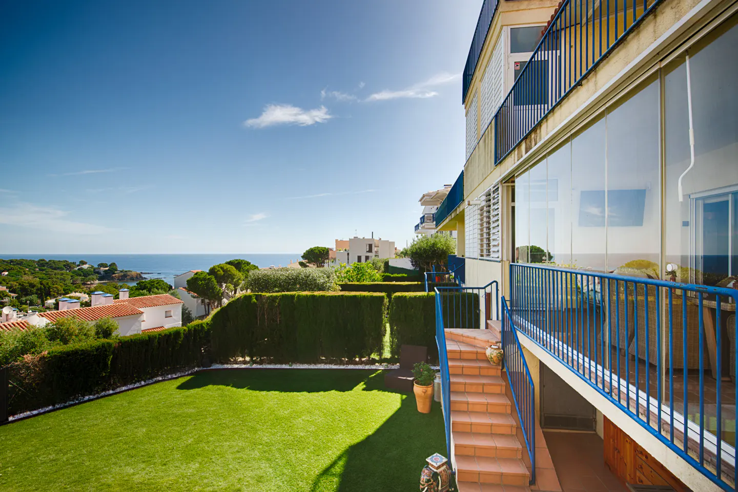 A sunny terrace with blue railings overlooks a green lawn, manicured hedges, and a distant blue sea under a clear sky.