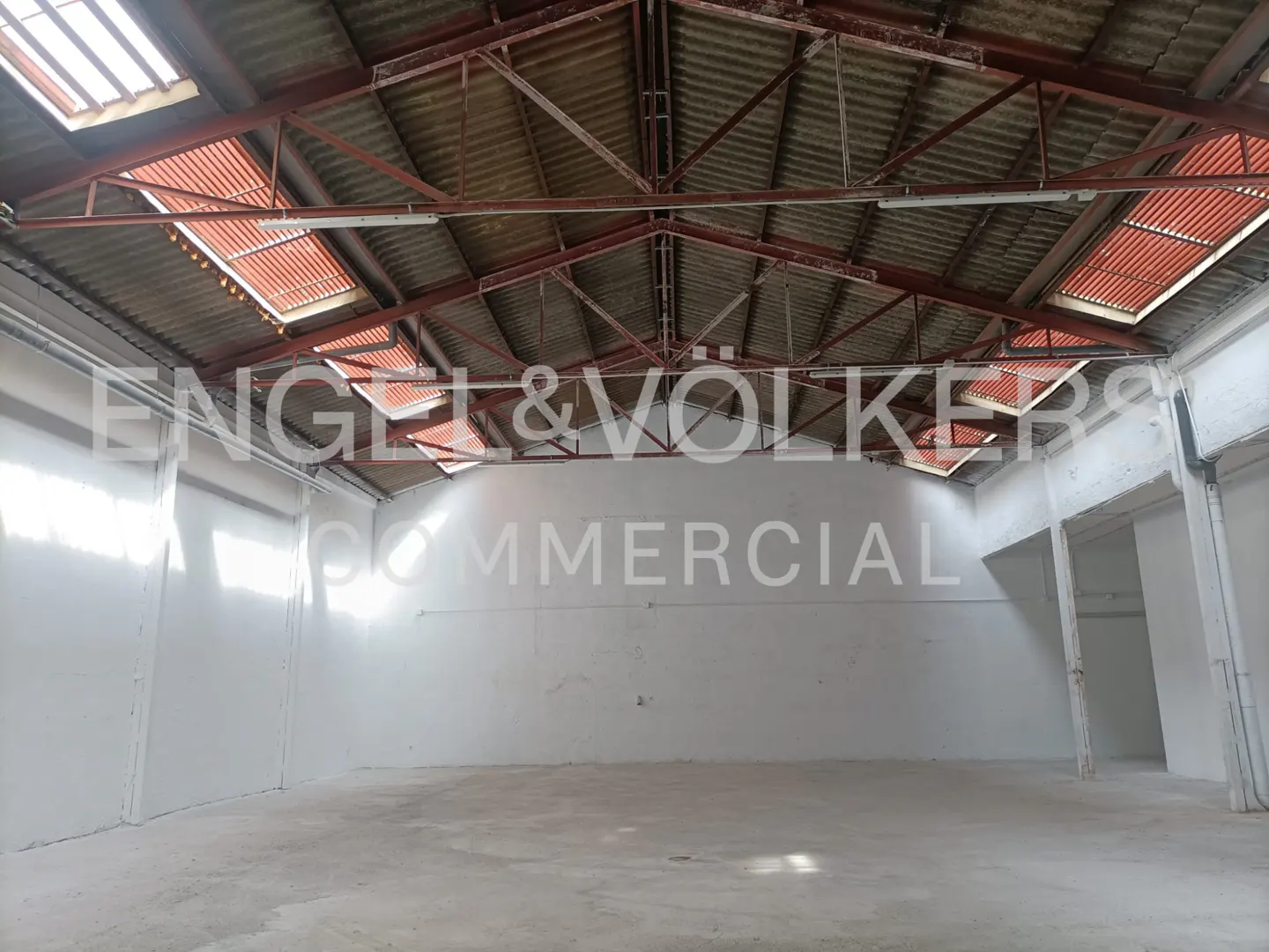 Empty warehouse interior with white walls, concrete floor, and red metal roof beams with skylights.
