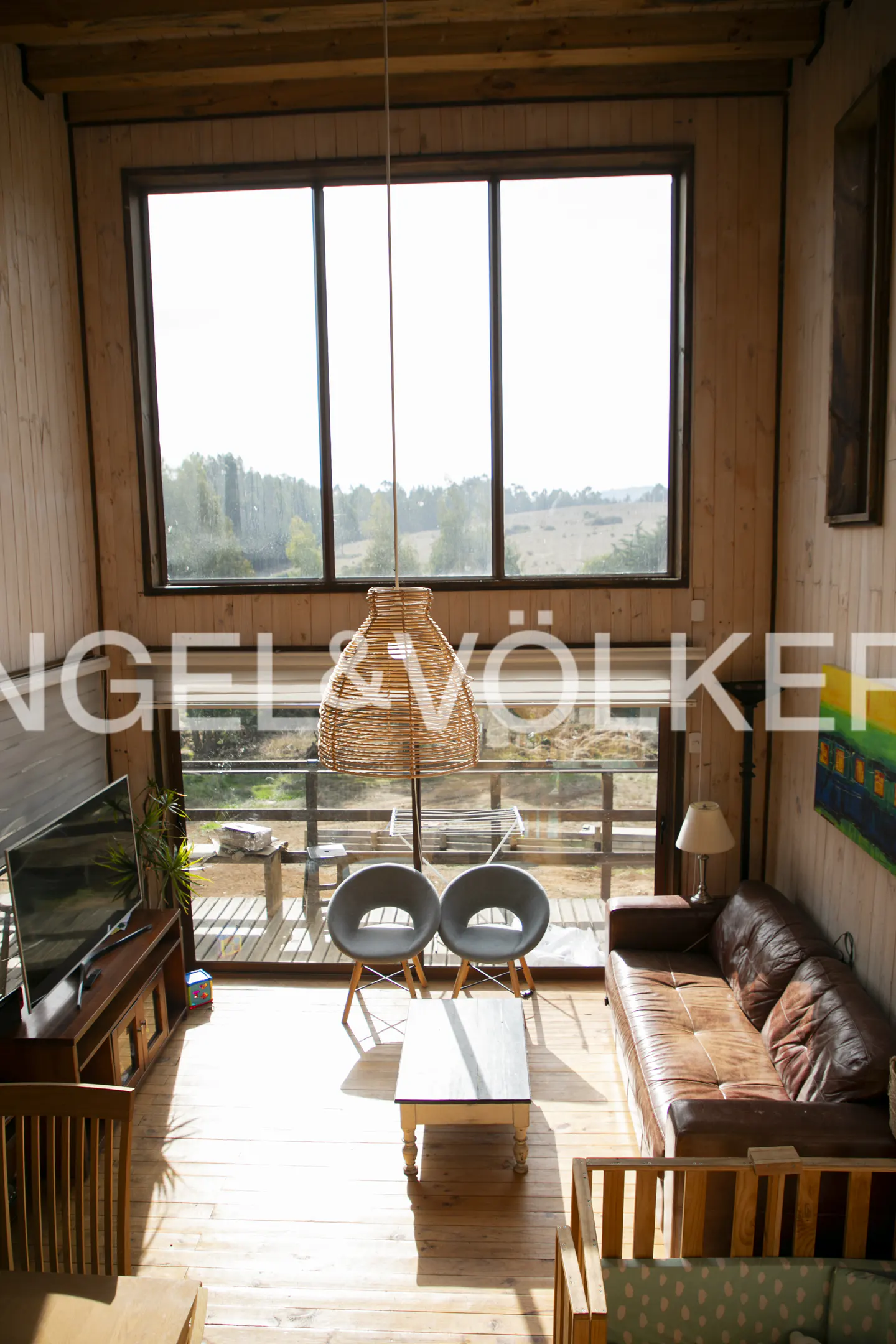 Living room with wood walls, large windows, brown leather sofa, two grey chairs, coffee table, and a woven pendant light.
