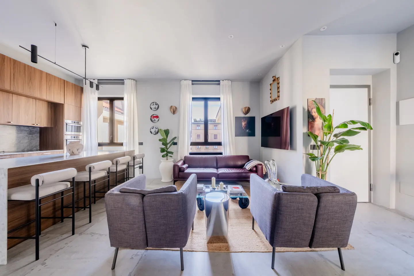 Modern open-plan living room with a wood kitchen, leather sofa, two grey armchairs, and a large potted plant.
