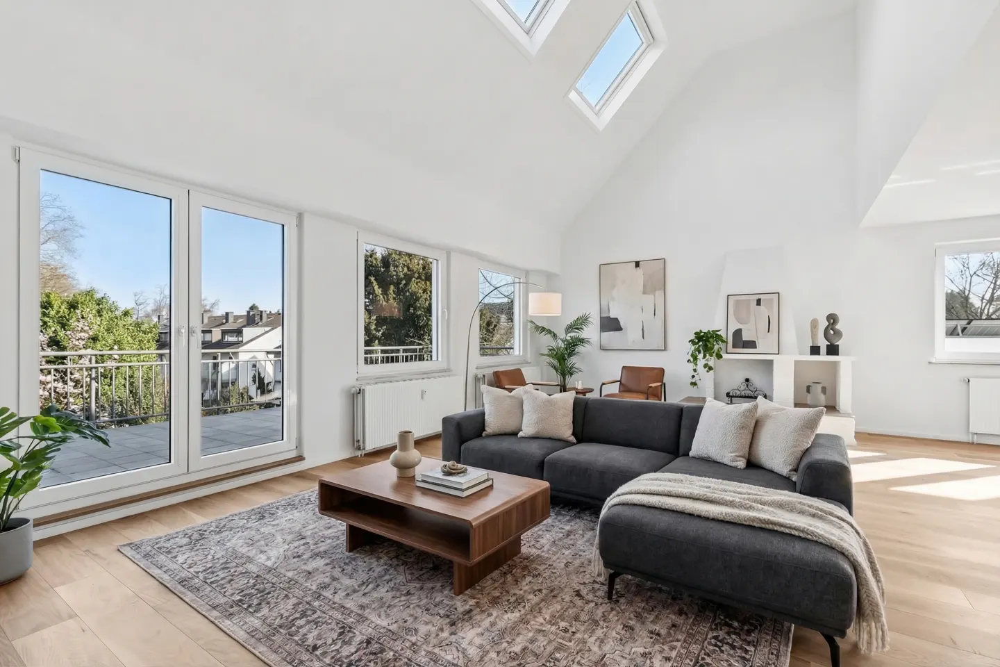 Bright living room with hardwood floors, a gray sectional sofa, and a wooden coffee table on a patterned rug. Balcony doors and skylights let in natural light.