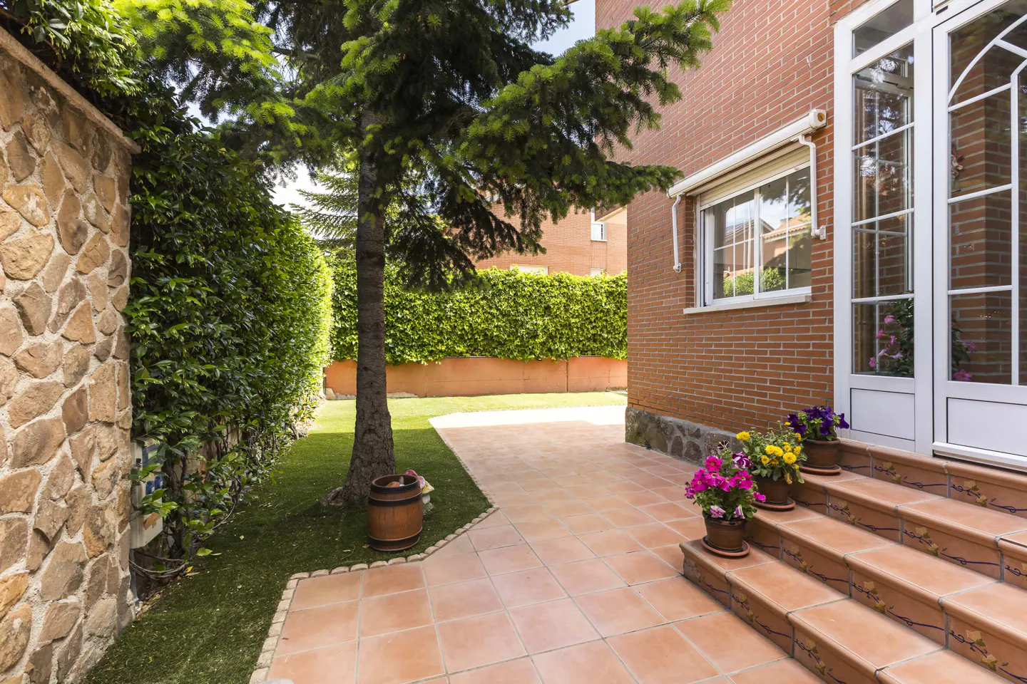 Brick house exterior with tiled patio, green lawn, and tall hedge. Potted flowers line the steps to a white-framed glass door.