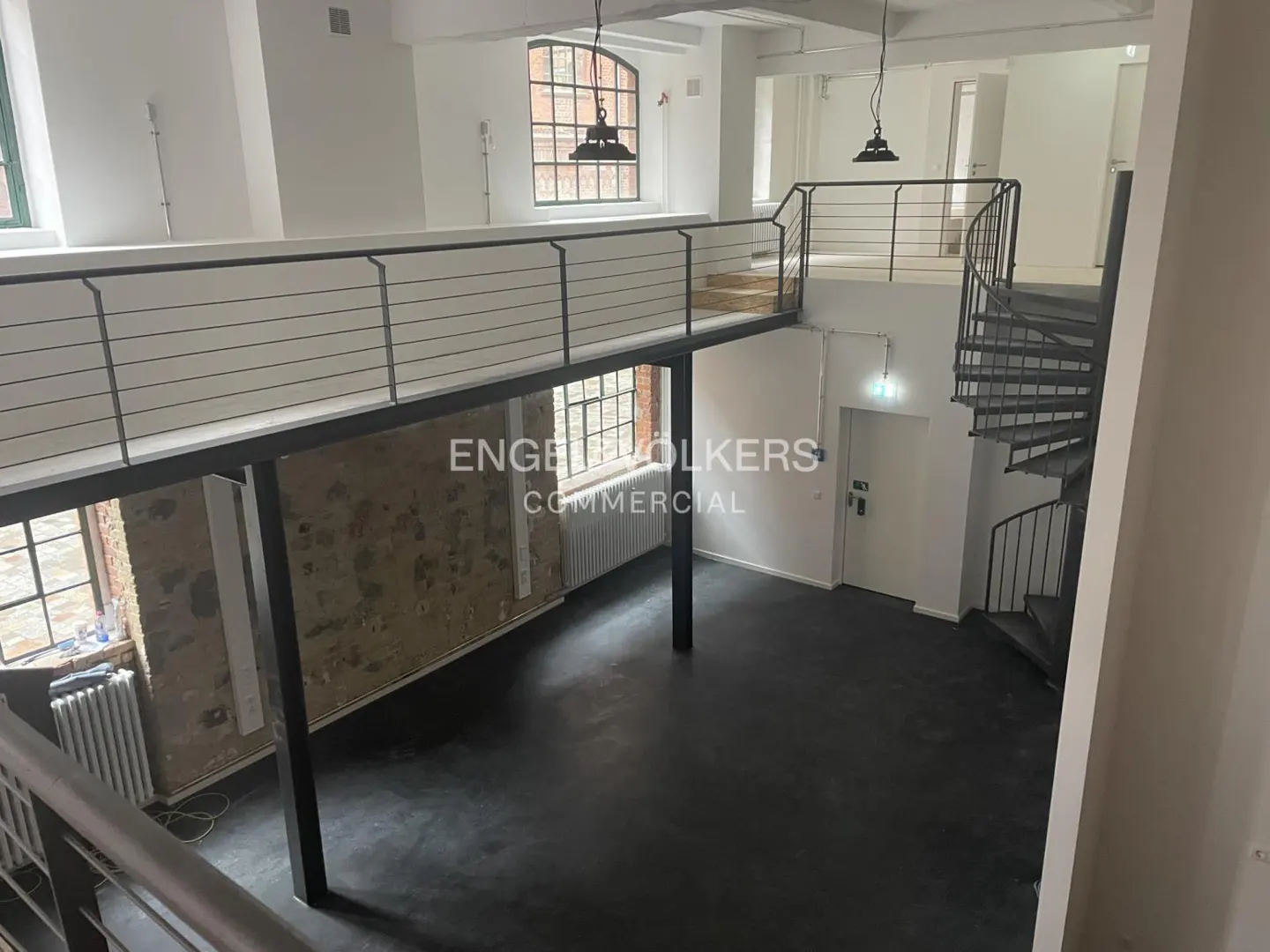 Interior view of a commercial space with a black floor, a mezzanine with metal railings, and a spiral staircase. Large windows and white walls provide natural light.