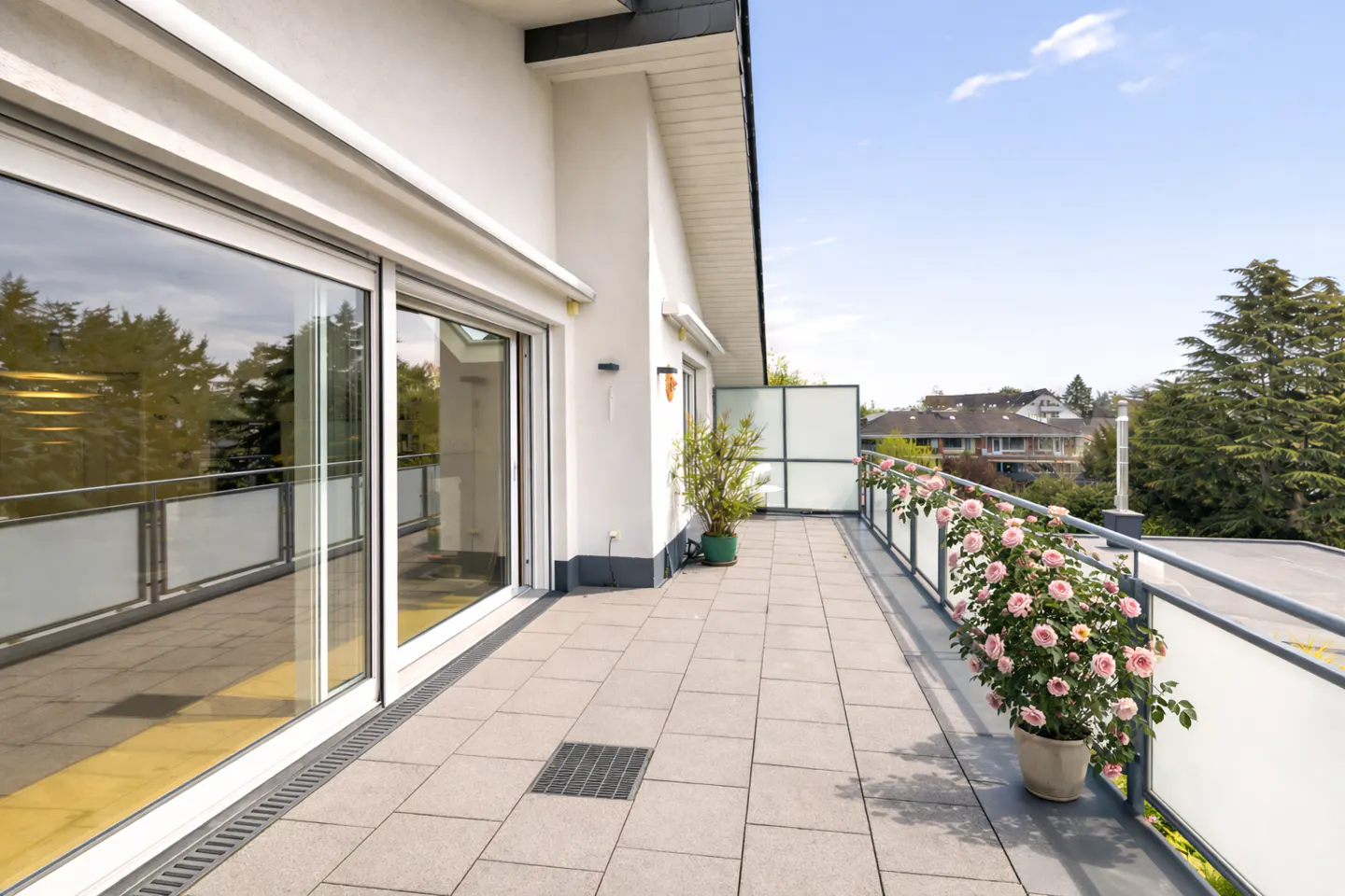 A long, gray-tiled balcony with a metal railing and pink roses in pots overlooks a neighborhood. Sliding glass doors lead into a white building.