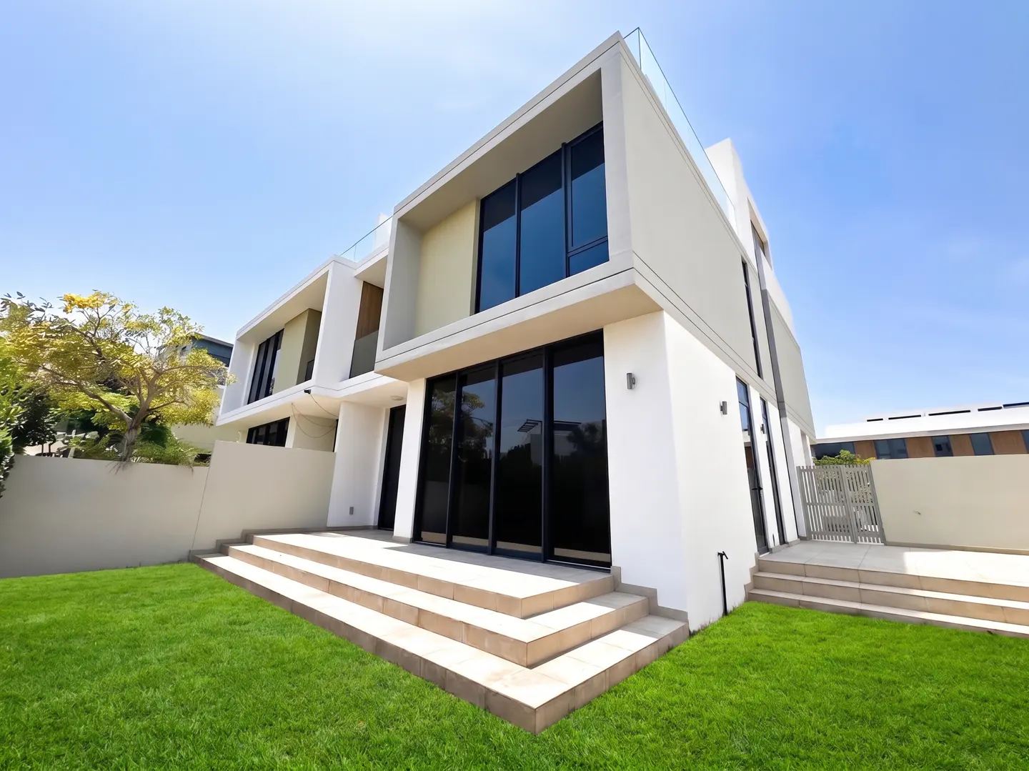 Modern two-story white home with large glass windows, stone patio steps, and a lush green lawn under a clear blue sky.