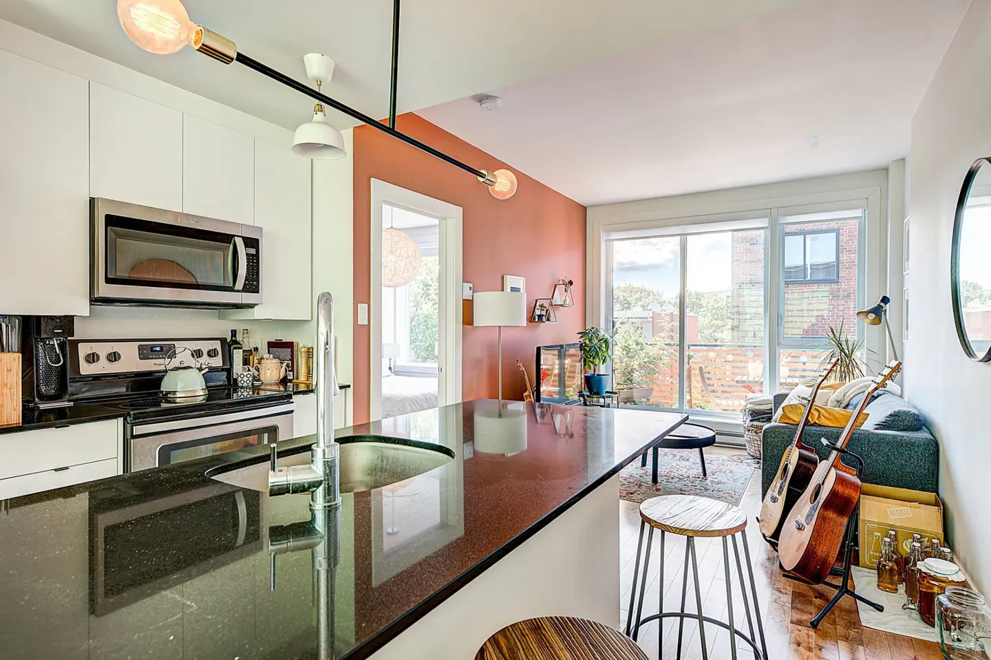 Bright, modern kitchen and living space with black granite island, white cabinets, and guitars leaning against a gray sofa near a large window.