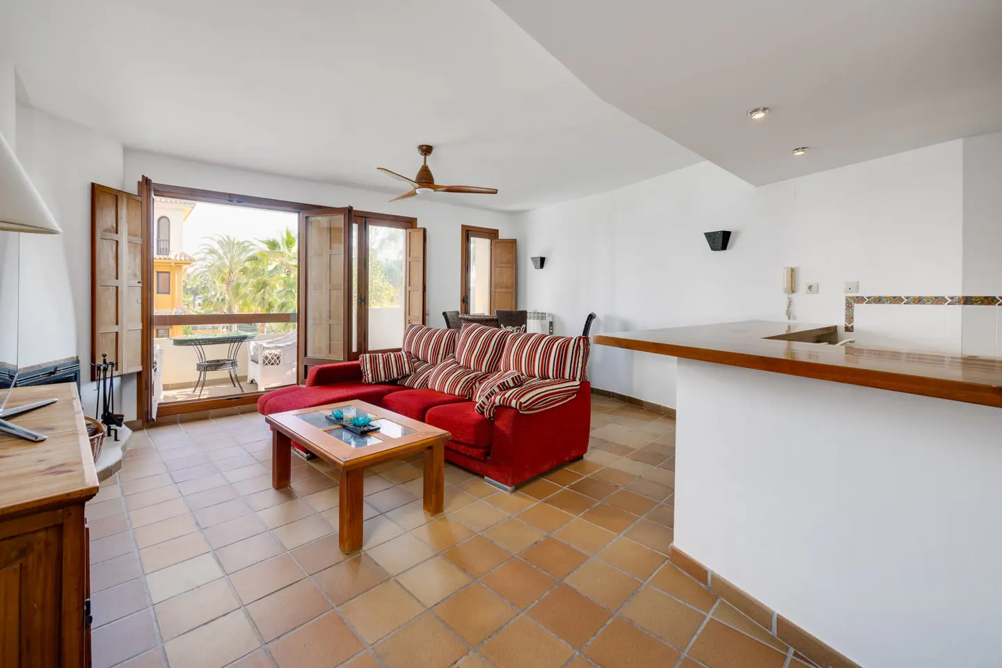 Bright living room with red sofa, wood table, and open balcony with palm trees. Tiled floor and white walls.