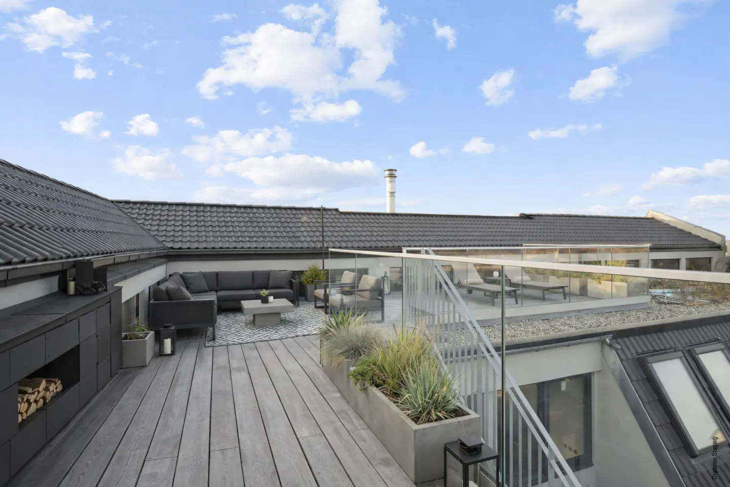 A rooftop patio with gray wood planks, a dark gray sectional sofa, and a glass railing under a blue sky.