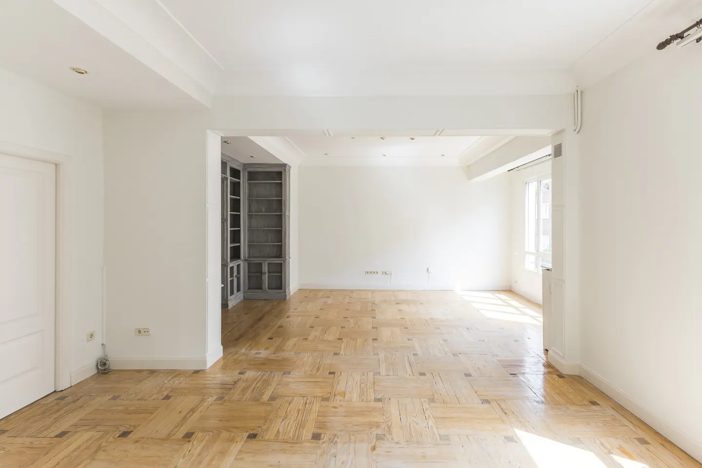 Bright, empty room with parquet floors and white walls. A gray built-in bookcase is visible in the background.