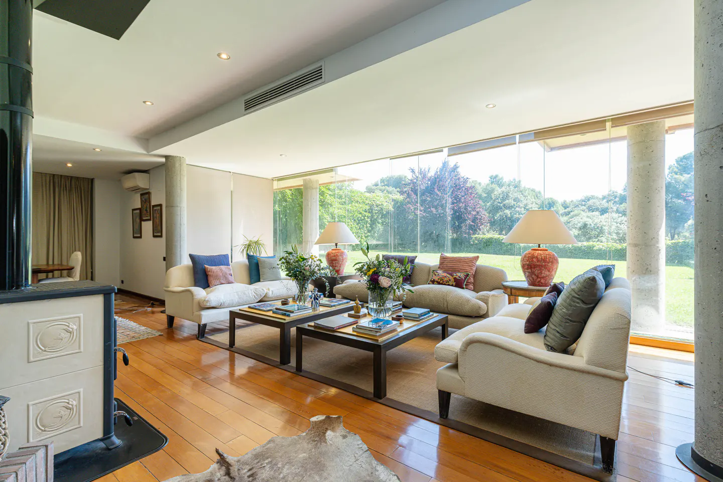 Bright living room with wood floors, white sofas, and large windows overlooking a green lawn. Two coffee tables sit in the center.