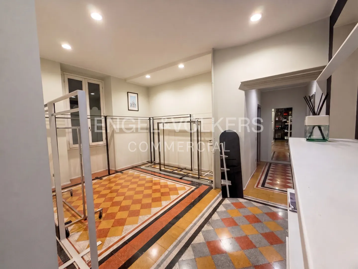 Empty retail space with white walls, metal clothing racks, and a patterned orange and grey diamond-tiled floor.