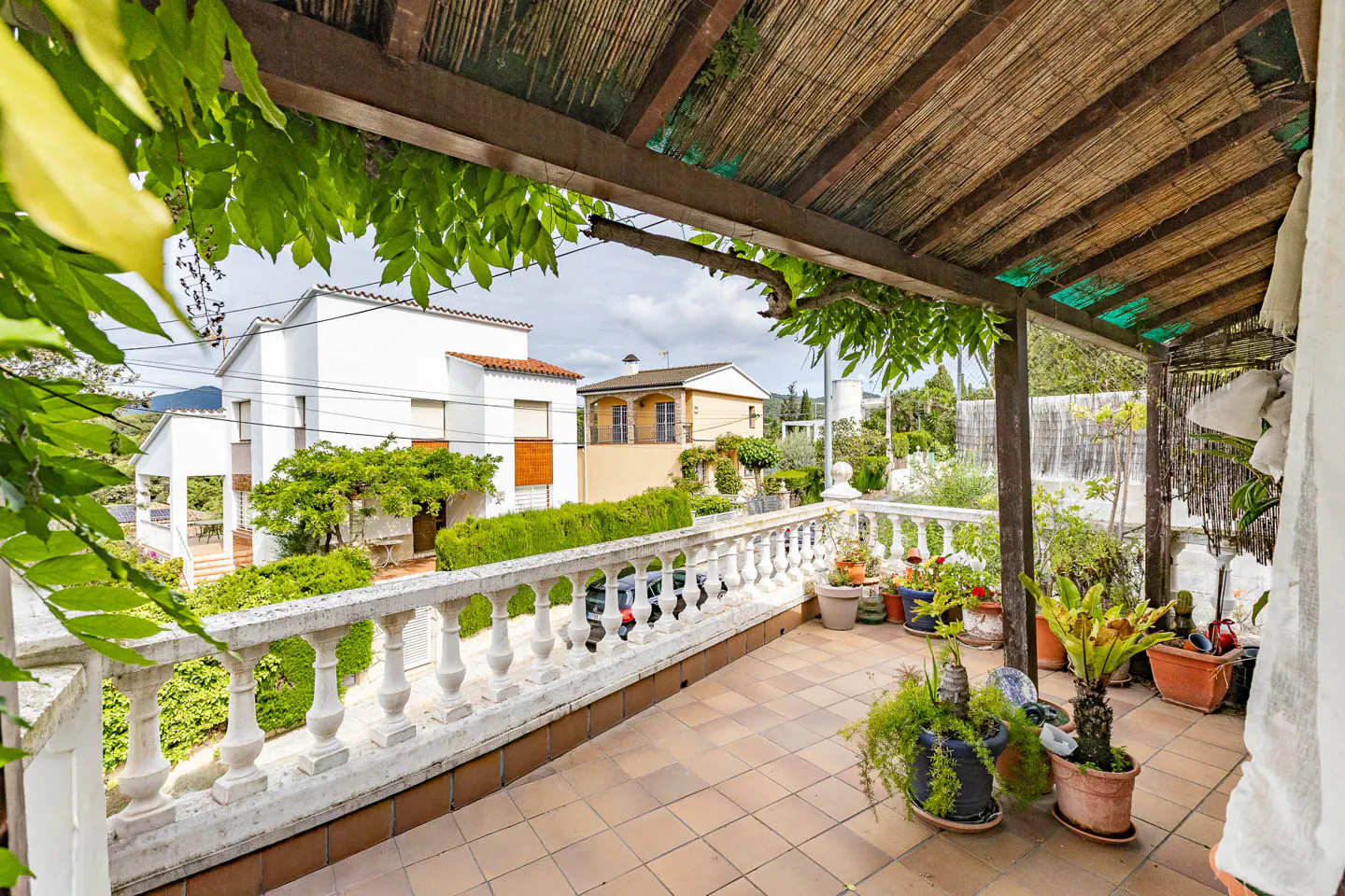 Covered patio with potted plants, a white balustrade, and a view of houses and greenery in the background.