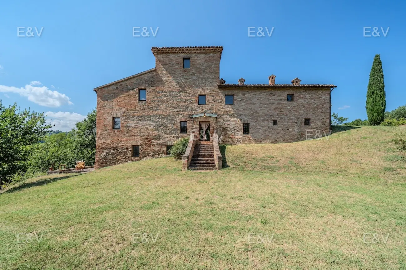 Exterior view of a two-story stone house with a tower, brick stairs, and a green lawn under a blue sky.