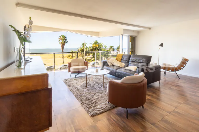 Bright living room with brown leather sofa, armchairs, and a view of the beach with palm trees.
