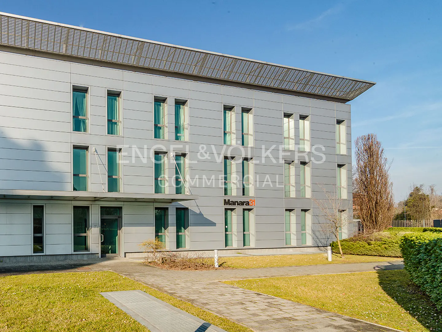 Modern three-story commercial building with silver metal panels, glass windows, and a paved walkway on a sunny day.