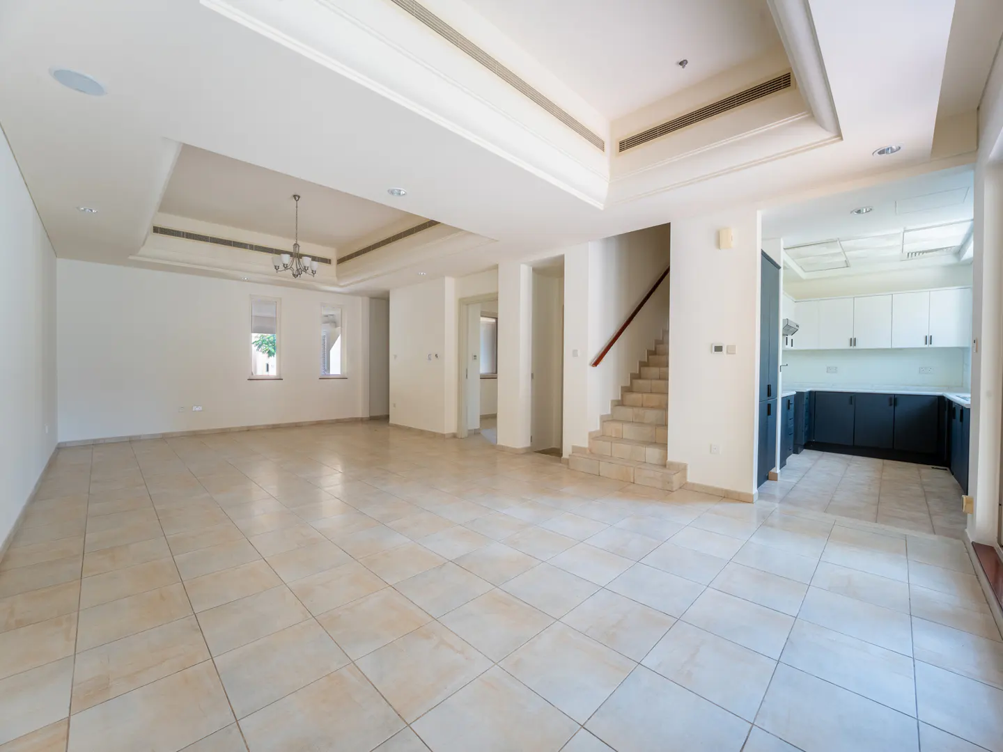 An interior shot of a large, empty living room with beige tile flooring, white walls, a staircase, and a view into the kitchen.