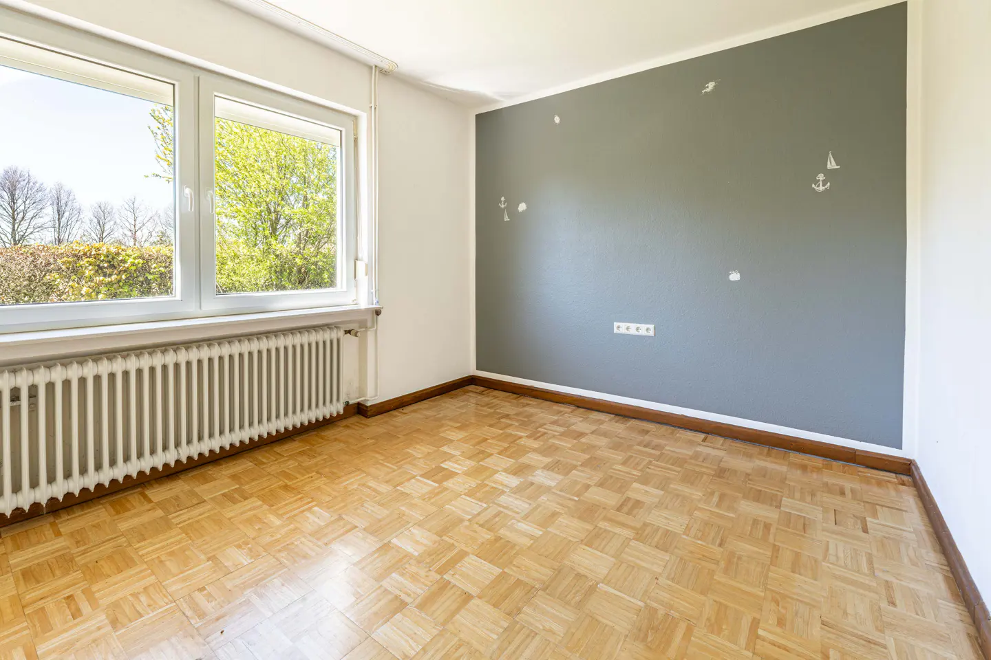 Empty room with parquet floor, radiator under a window, and a gray wall with nautical-themed decals.