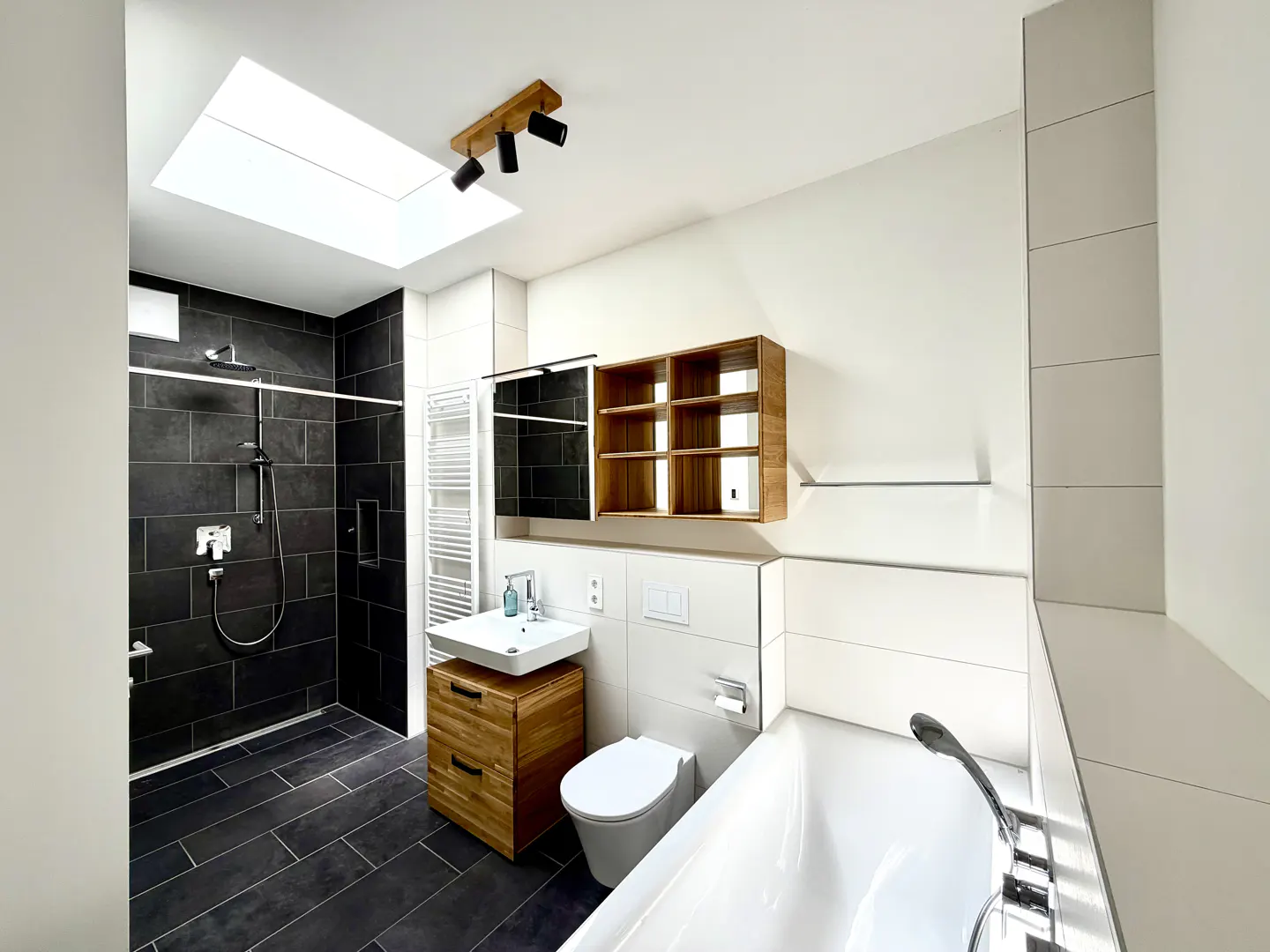 Bright bathroom with black tiled shower, white walls, and a skylight. A wooden vanity and shelf add warmth to the modern design.