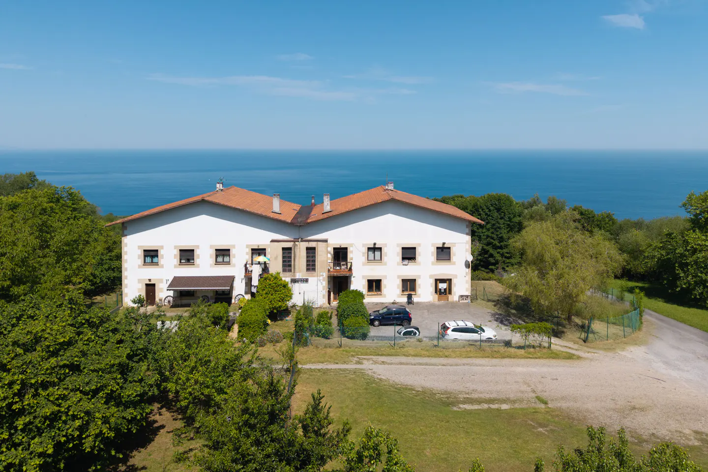Two-story white house with a red tile roof, surrounded by green trees, with a view of the blue ocean in the background.