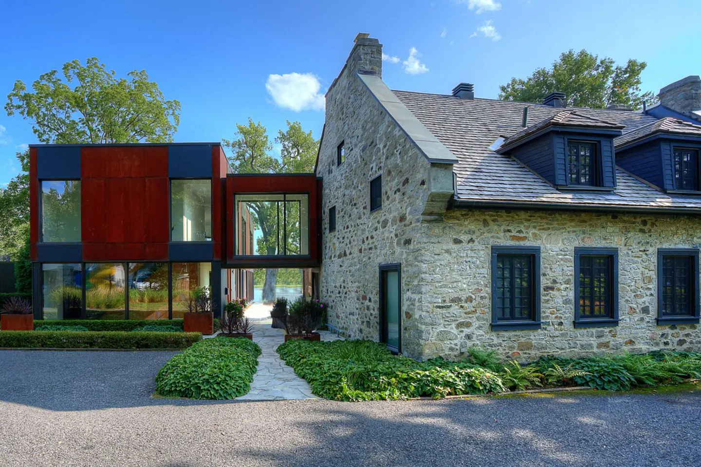 Exterior view of a stone house connected to a modern rust-colored building by a glass walkway, under a blue sky.