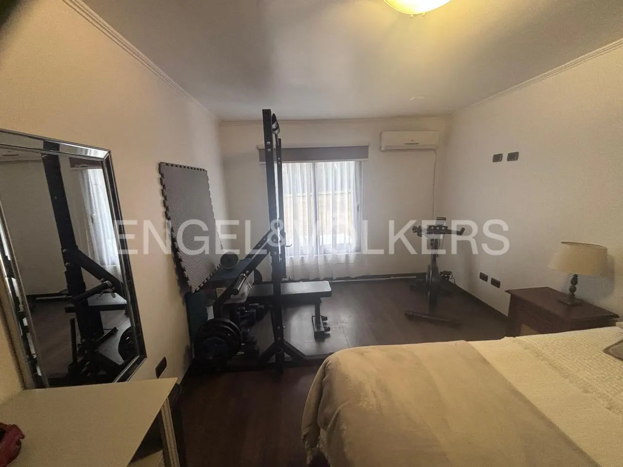 Home gym in a bedroom with a weight rack, exercise bike, mirror, and a bed with white bedding.