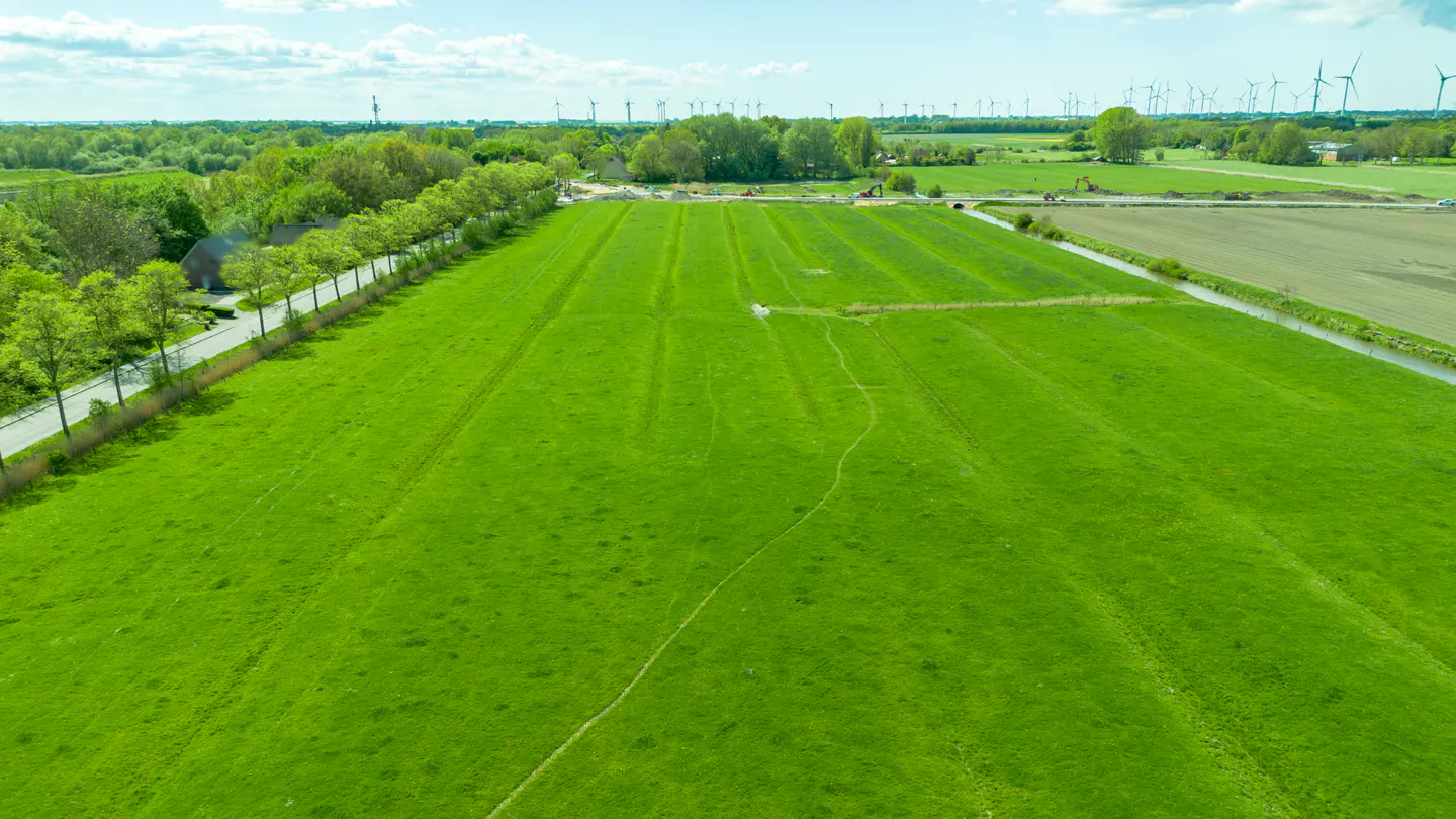Aerial view of a lush green field with trees, a road, and windmills in the distance under a blue sky.