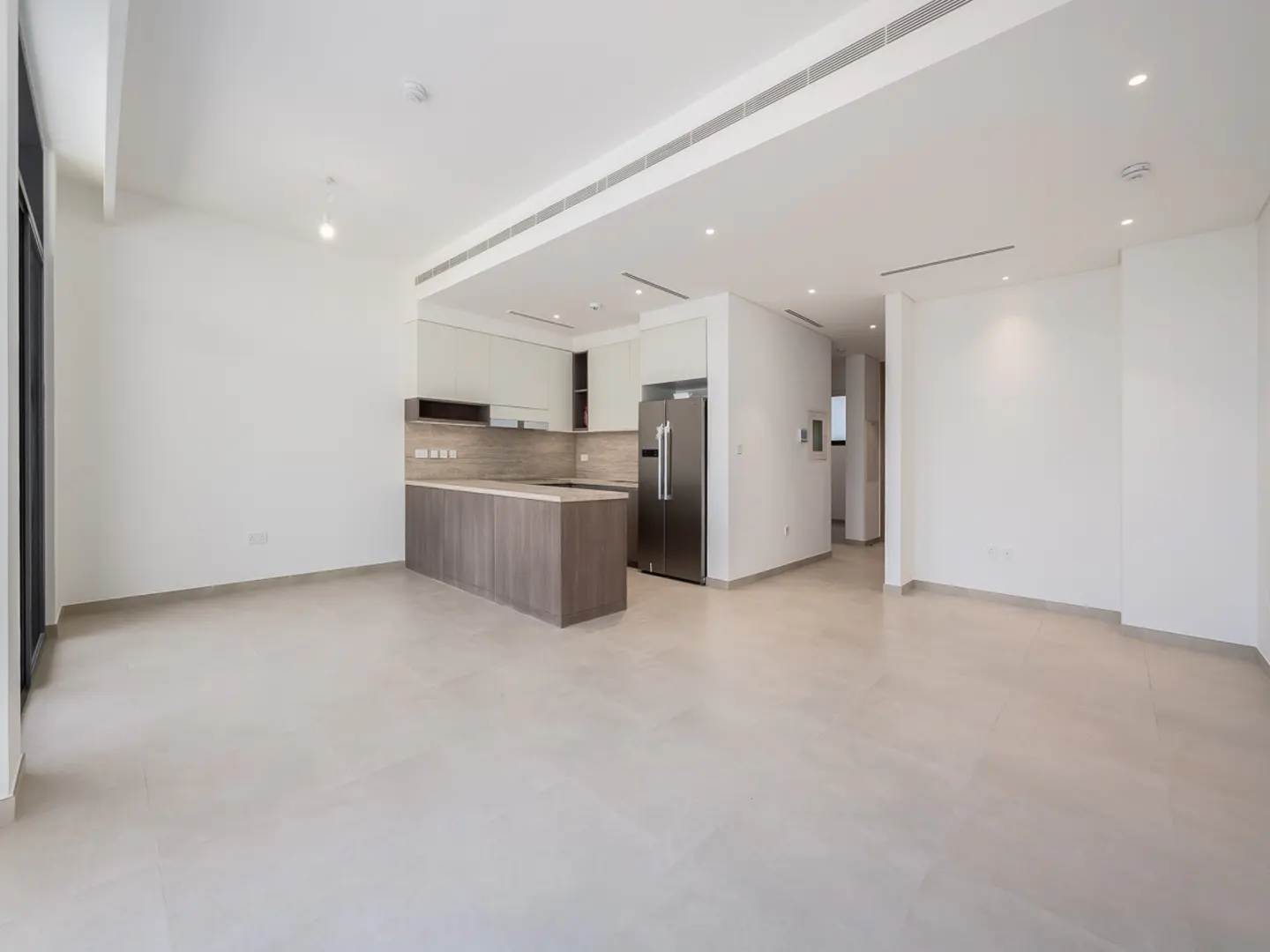 Bright, empty room with white walls and light tile floor. Kitchen area with island, stainless steel fridge, and light wood cabinets.